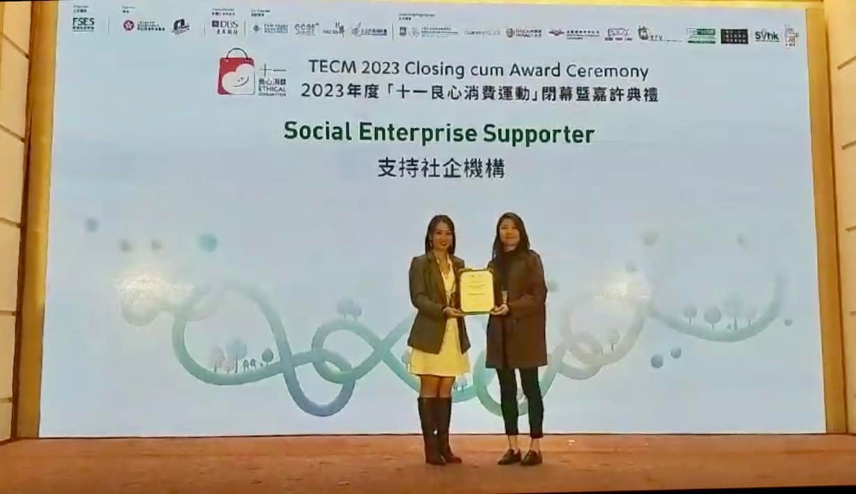 Two women on stage, one in a grey blazer, receives an award certificate from another woman at a ceremony.