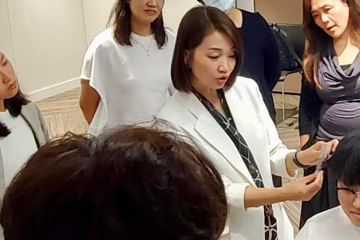 A female trainer in a white blazer actively explains a concept, gesturing with her hands to a group of attentive participants.
