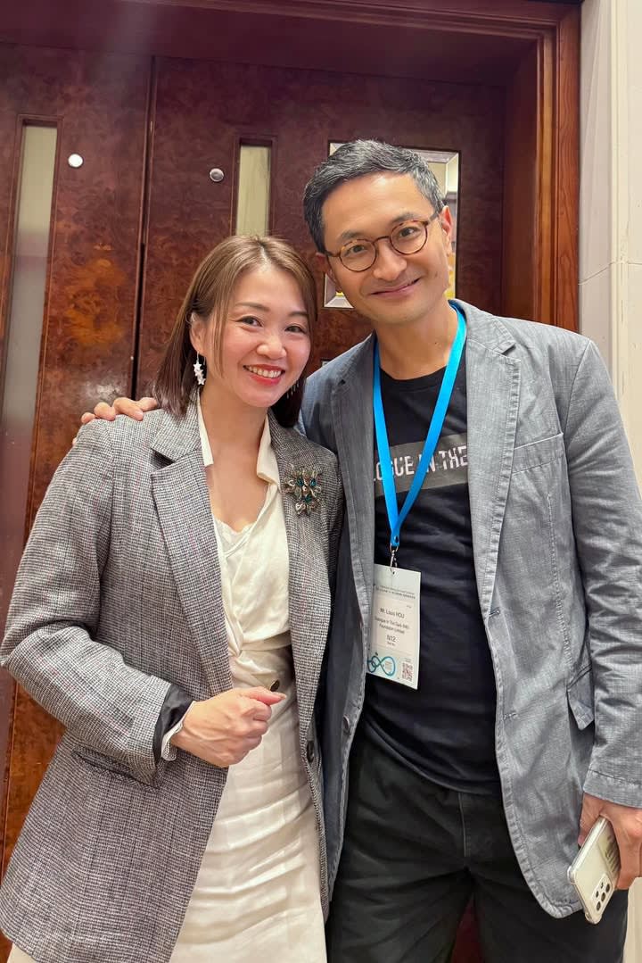 A smiling woman in a grey blazer and a man in glasses pose for a photo indoors, looking friendly.