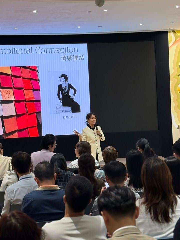 A female trainer speaks into a microphone on stage, with a screen showing "Emotional Connection" behind her and an audience.