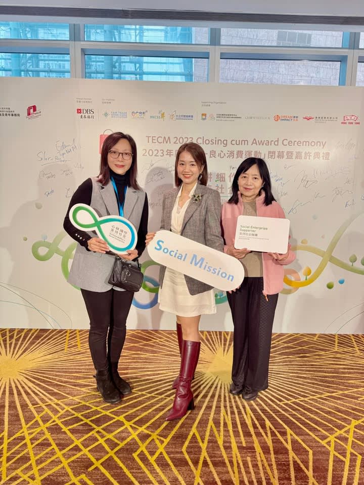 Three smiling women hold signs for social enterprises at an event, standing in front of a branded backdrop.