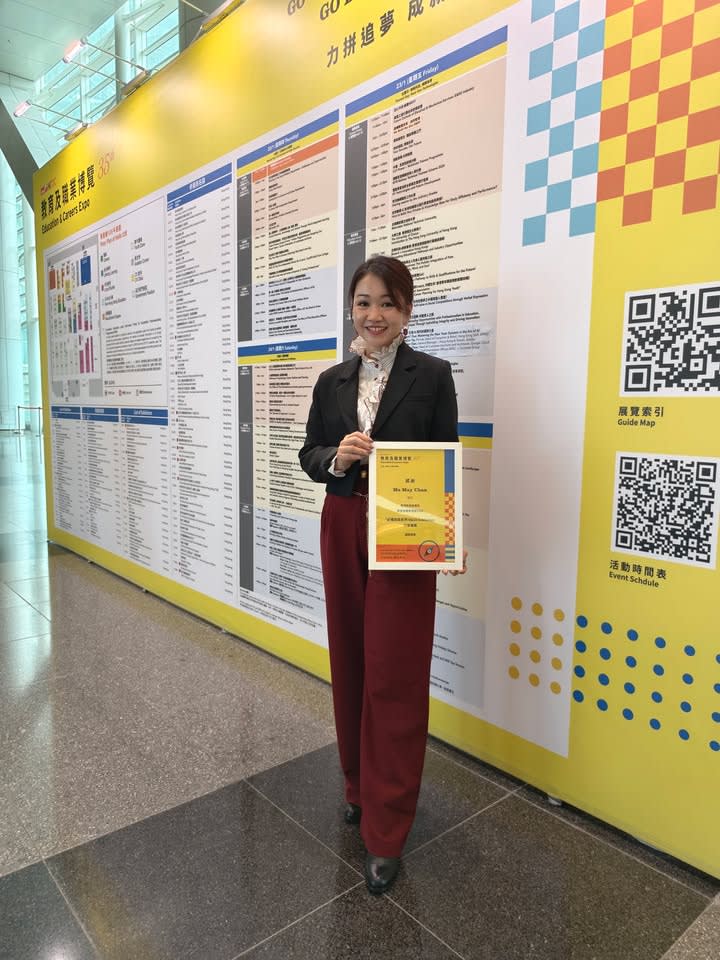 A smiling trainer in a hallway, holding a certificate, standing next to a large event information board.