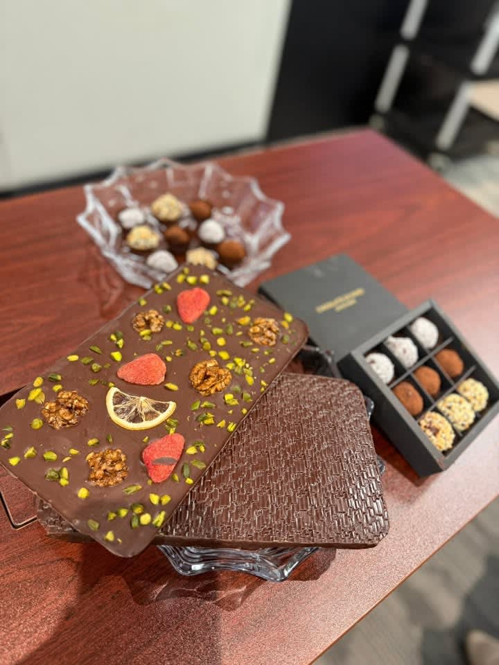 An assortment of chocolates on a wooden table, including a large bar with nuts and fruit, textured bars, and truffles.