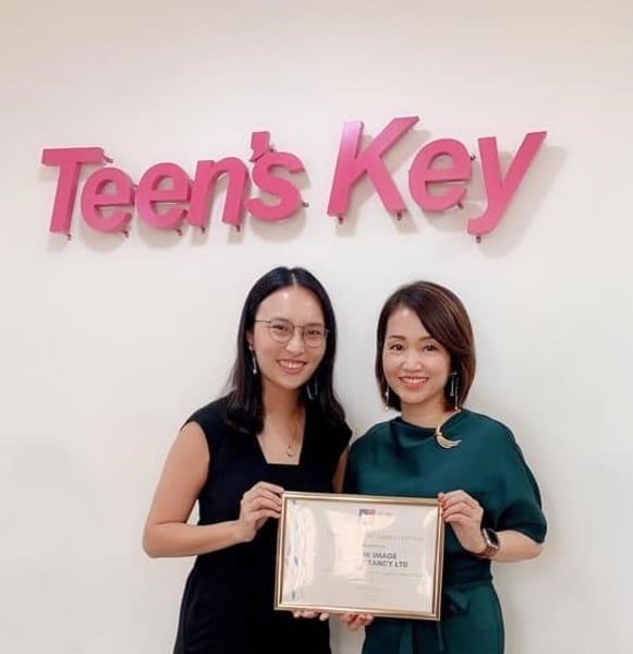 Two smiling women hold a framed certificate in front of a wall with pink 'Teen's Key' letters.