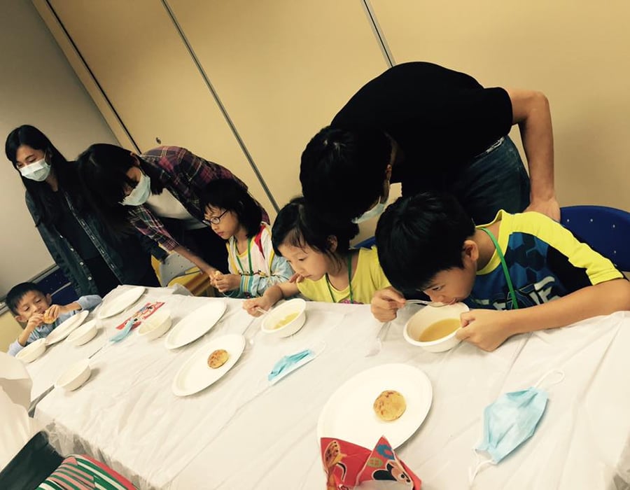 Children and adults at a long table. Three children are eating soup from bowls, while adults supervise them.