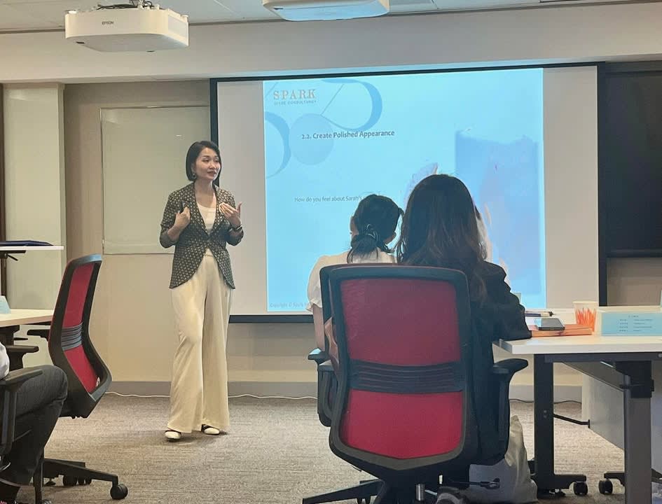 Female trainer gestures while presenting to two attendees in a modern training room with a projector screen showing slides.