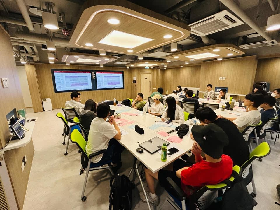Participants are seated around large tables in a modern training room, engaged in a session with screens displaying content.