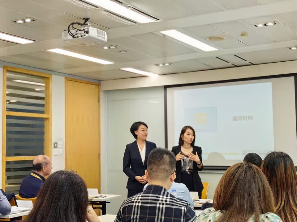 Two women trainers presenting to an audience in a training room, with a projector screen behind them.