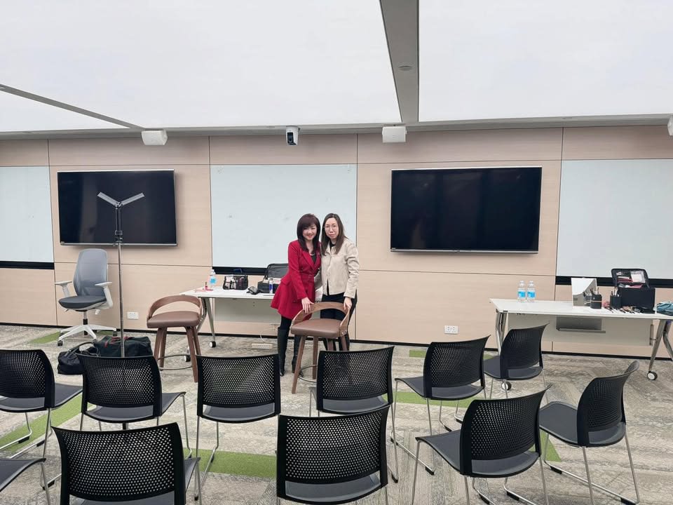 Two women, one in red and one in beige, stand smiling in a training room with chairs, whiteboards, and monitors.