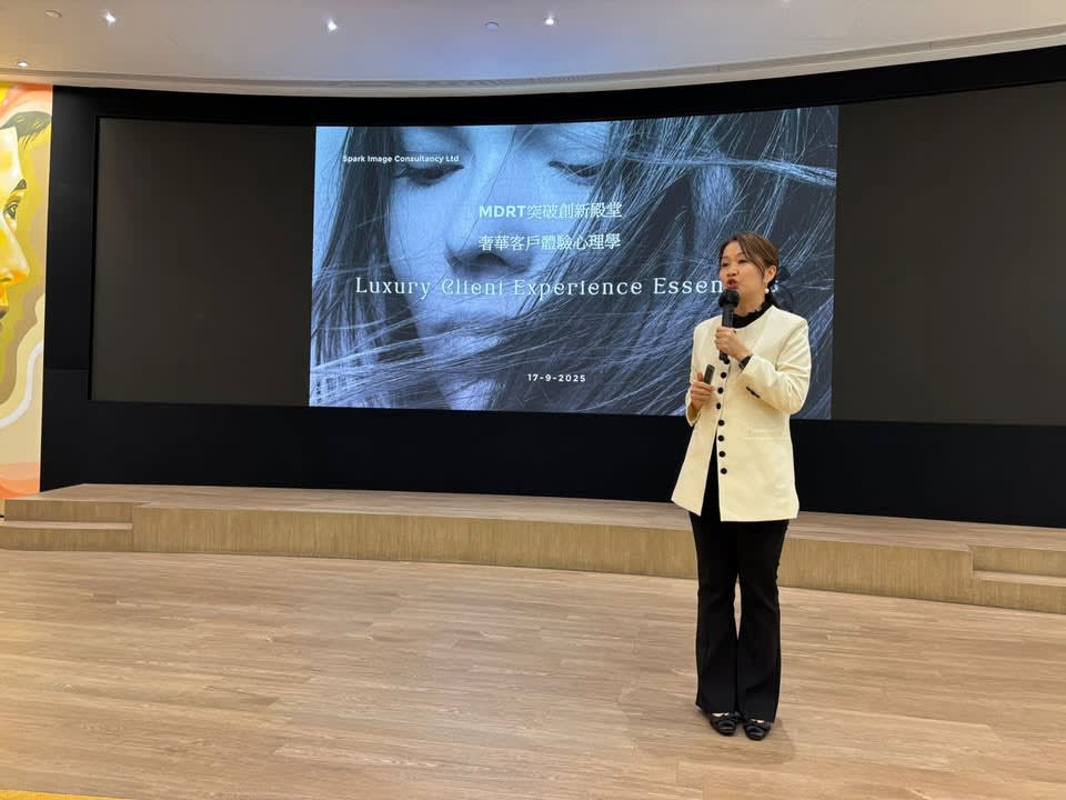A female trainer stands on a stage holding a microphone, with a large screen displaying "Spark Image Consultancy" and event details.